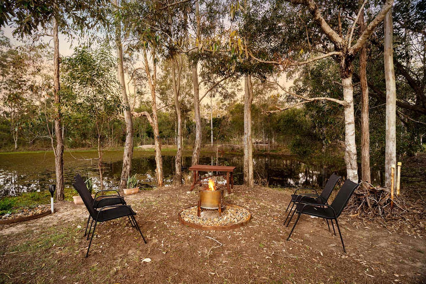 An outdoor seating area with two chairs, a table, and a fire pit surrounded by trees and a body of water.