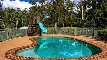 A swimming pool with a slide, surrounded by a brick border and a metal fence.