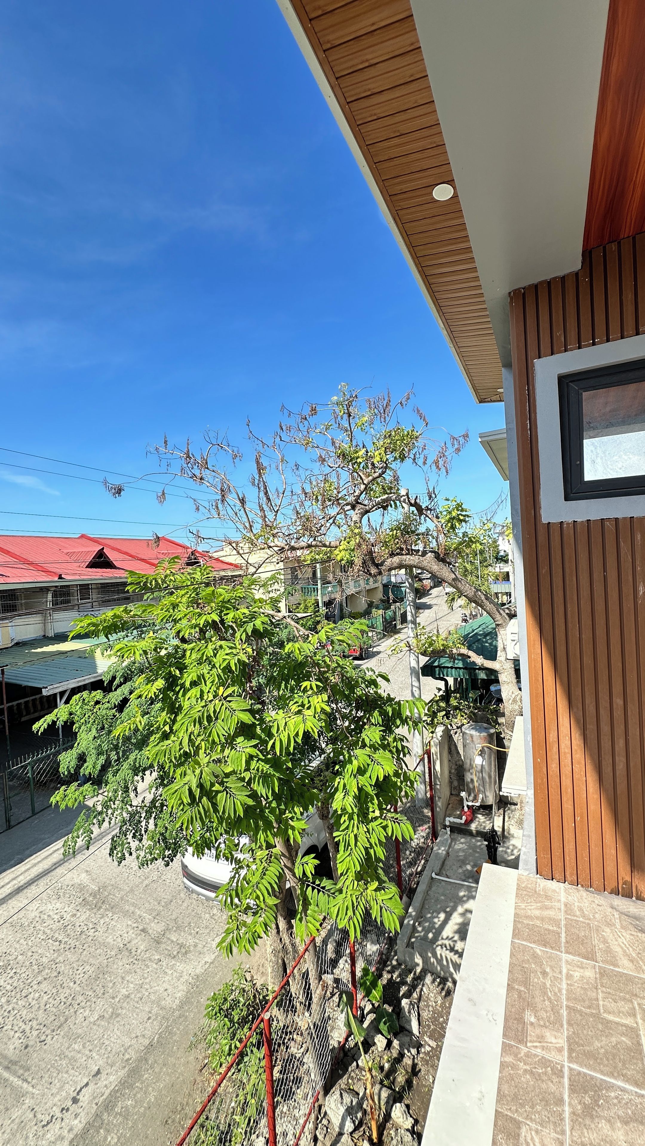 A balcony with a view of a red-roofed building, a tree, and a stone wall.