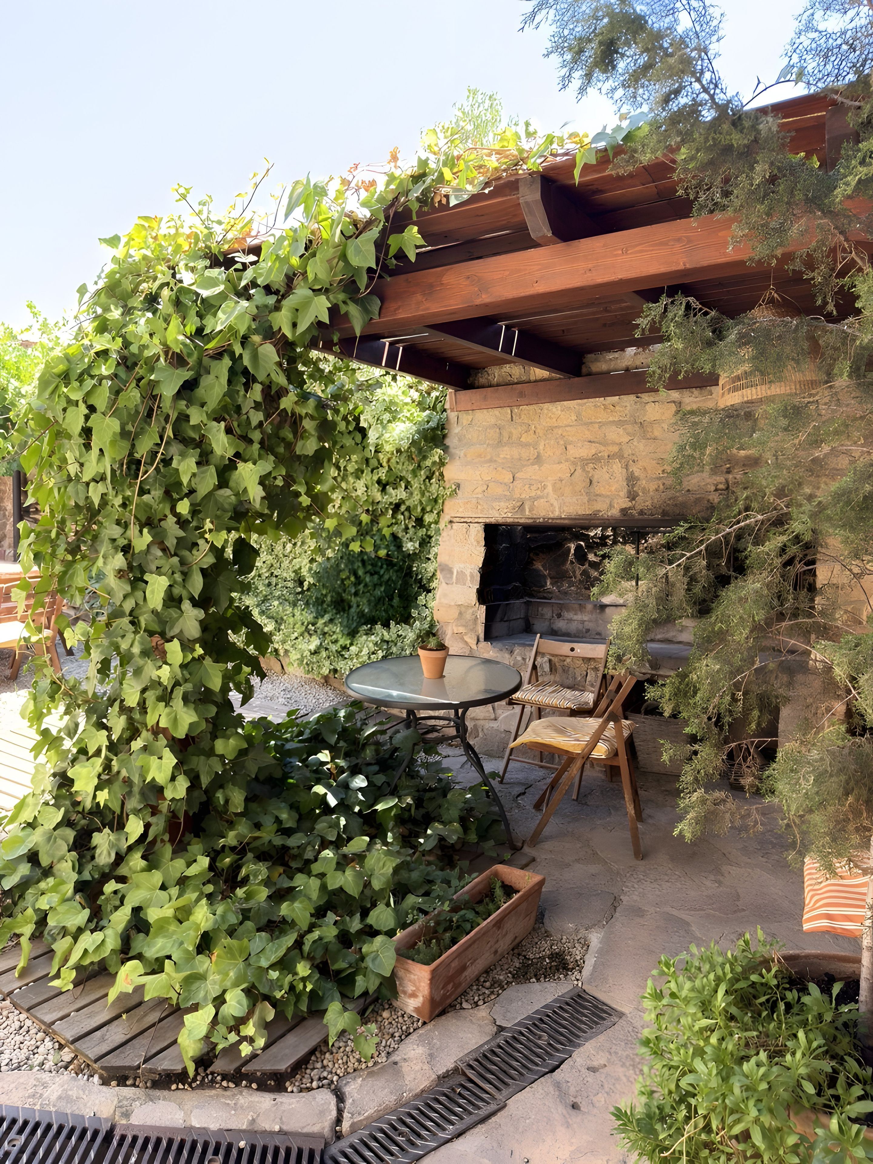 Une terrasse extérieure dotée d’une cheminée en pierre, de meubles en bois et d’une table entourée de verdure.