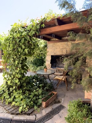 An outdoor patio with a stone fireplace, wooden furniture, and a table surrounded by greenery.