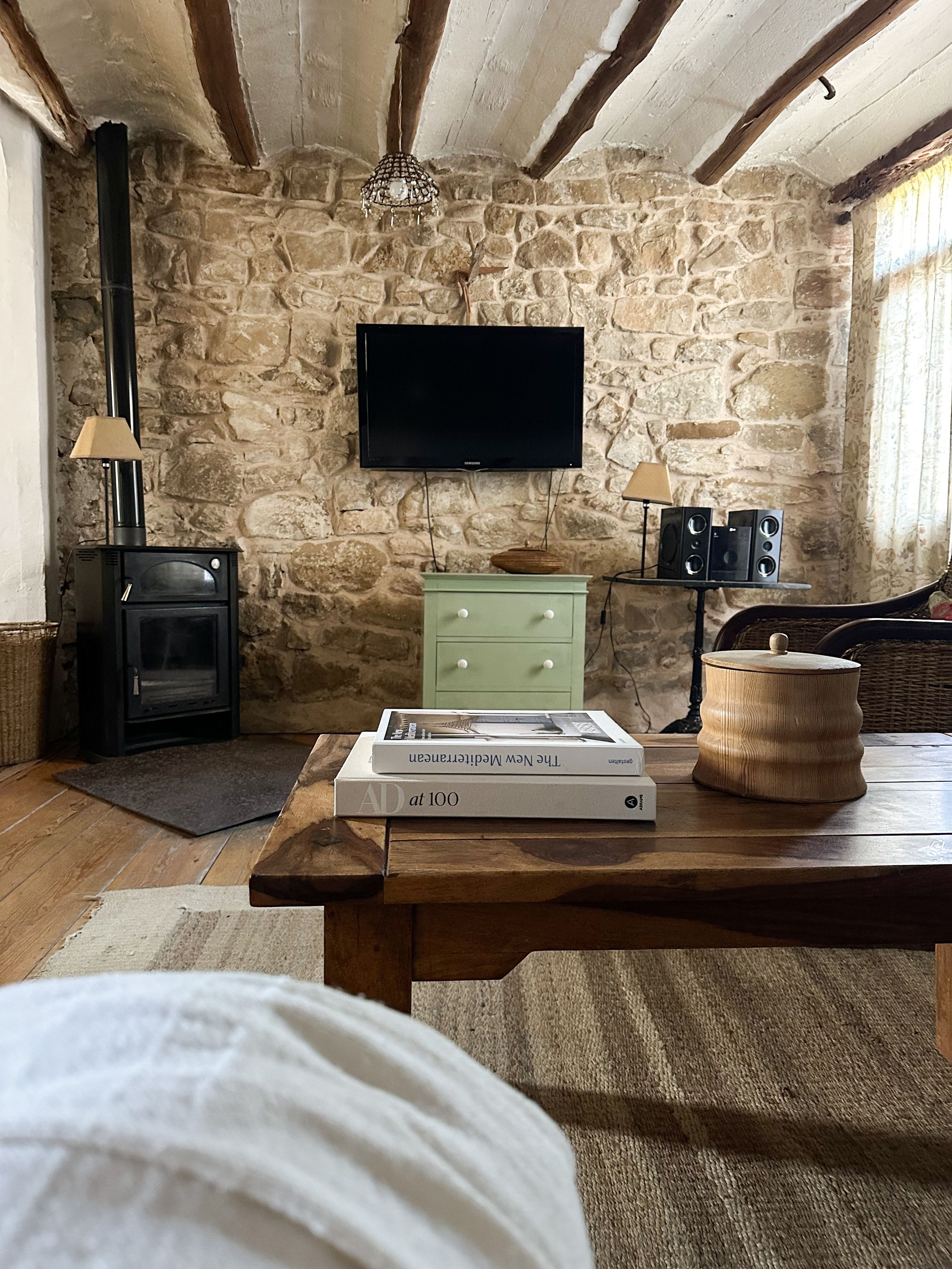 Une pièce chaleureuse avec un mur en pierre, une cheminée, une télévision et une table sur laquelle sont posés des livres et une lampe.