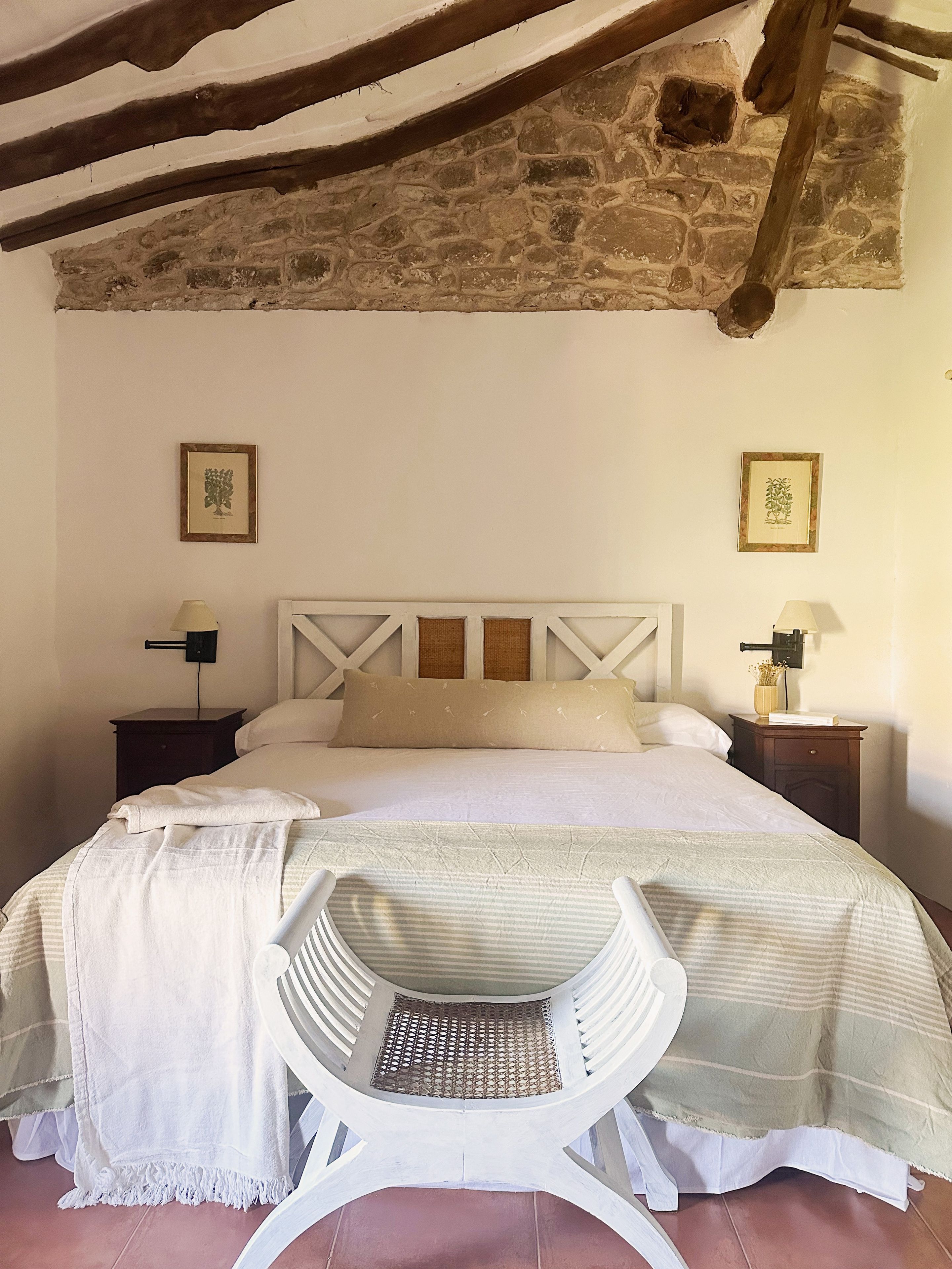 Een slaapkamer met een stenen muur, een bed met een witte hoofdbord, twee nachtkastjes met lampen, en een stoel voor het bed.