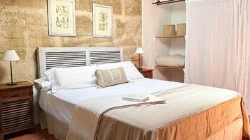 Un dormitorio con techo de madera, una cama con ropa de cama blanca, dos cuadros enmarcados en la pared, una mesita de noche con una lámpara y una ventana con cortinas.
