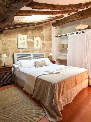 A bedroom with a wooden ceiling, a bed with white bedding, two framed pictures on the wall, a bedside table with a lamp, and a window with curtains.