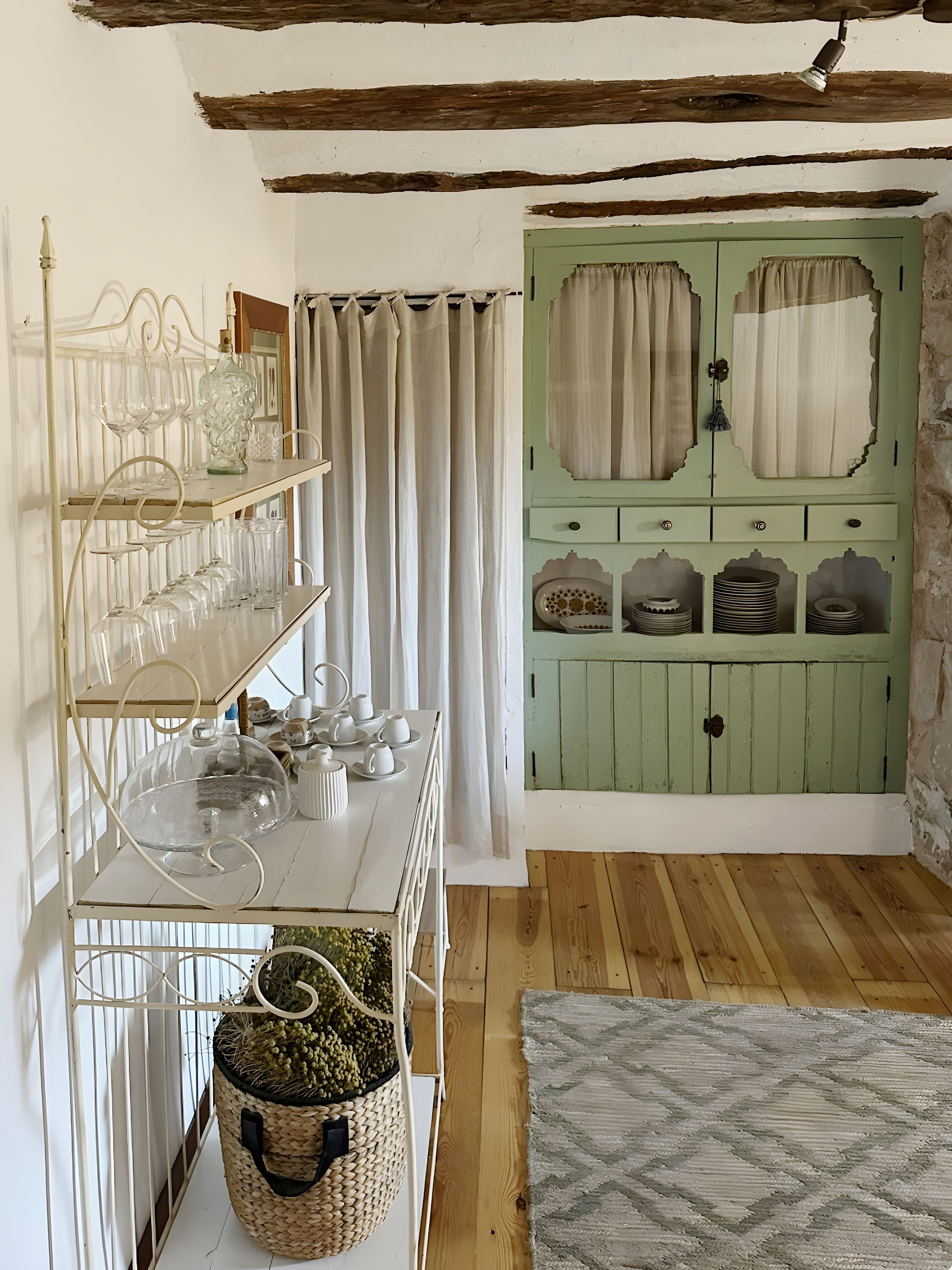 Una habitación con suelo de madera, una puerta verde con cortinas, un estante blanco con cristalería y una cesta tejida.