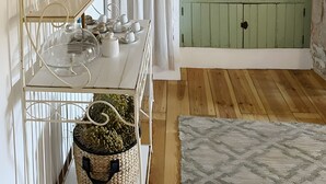 Quarto com piso de madeira, porta verde com cortinas, prateleira branca com copos e cesta de palha.