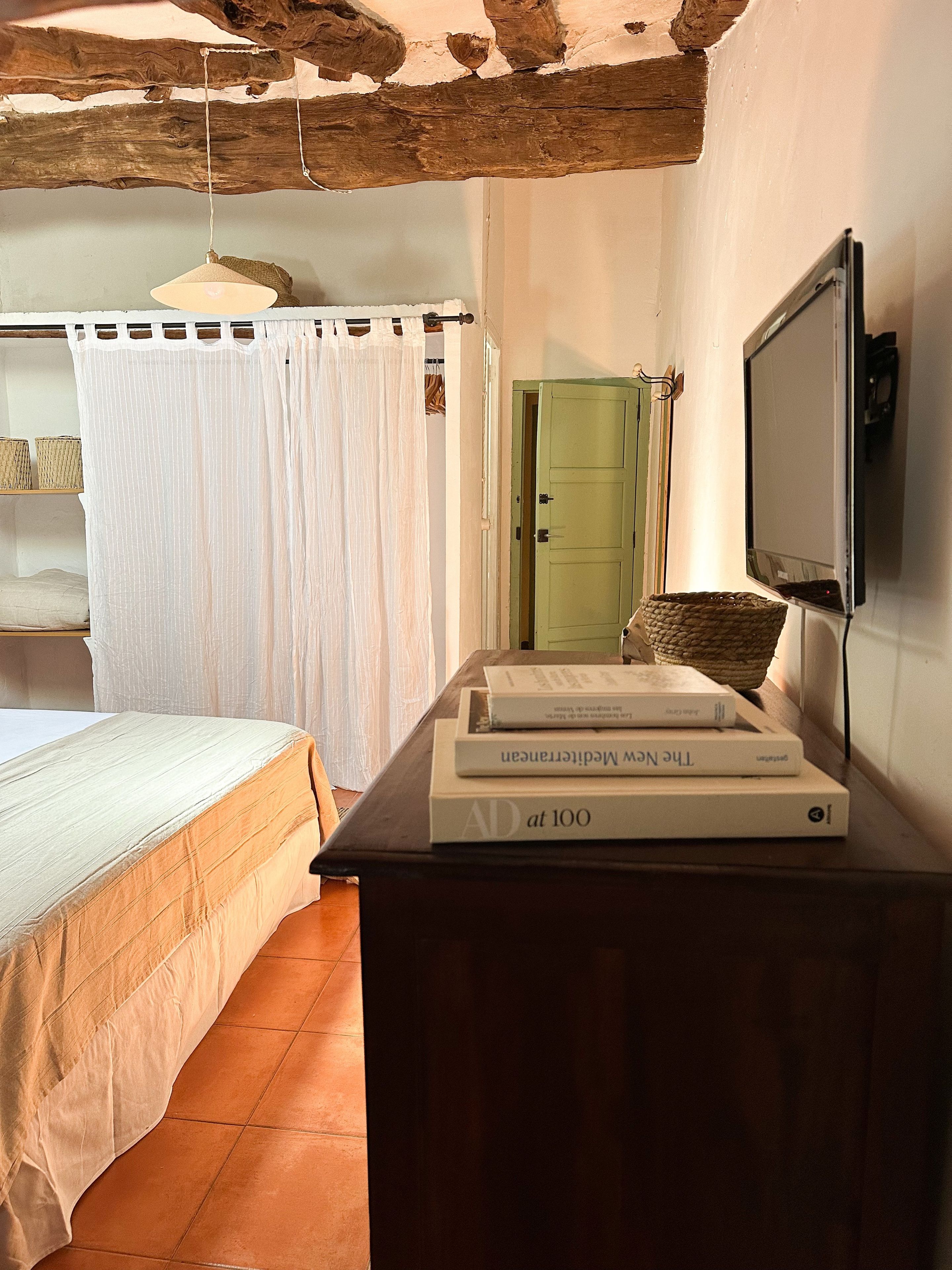 Un dormitorio con una cama, una mesa de madera con libros, una puerta verde y una cortina blanca.