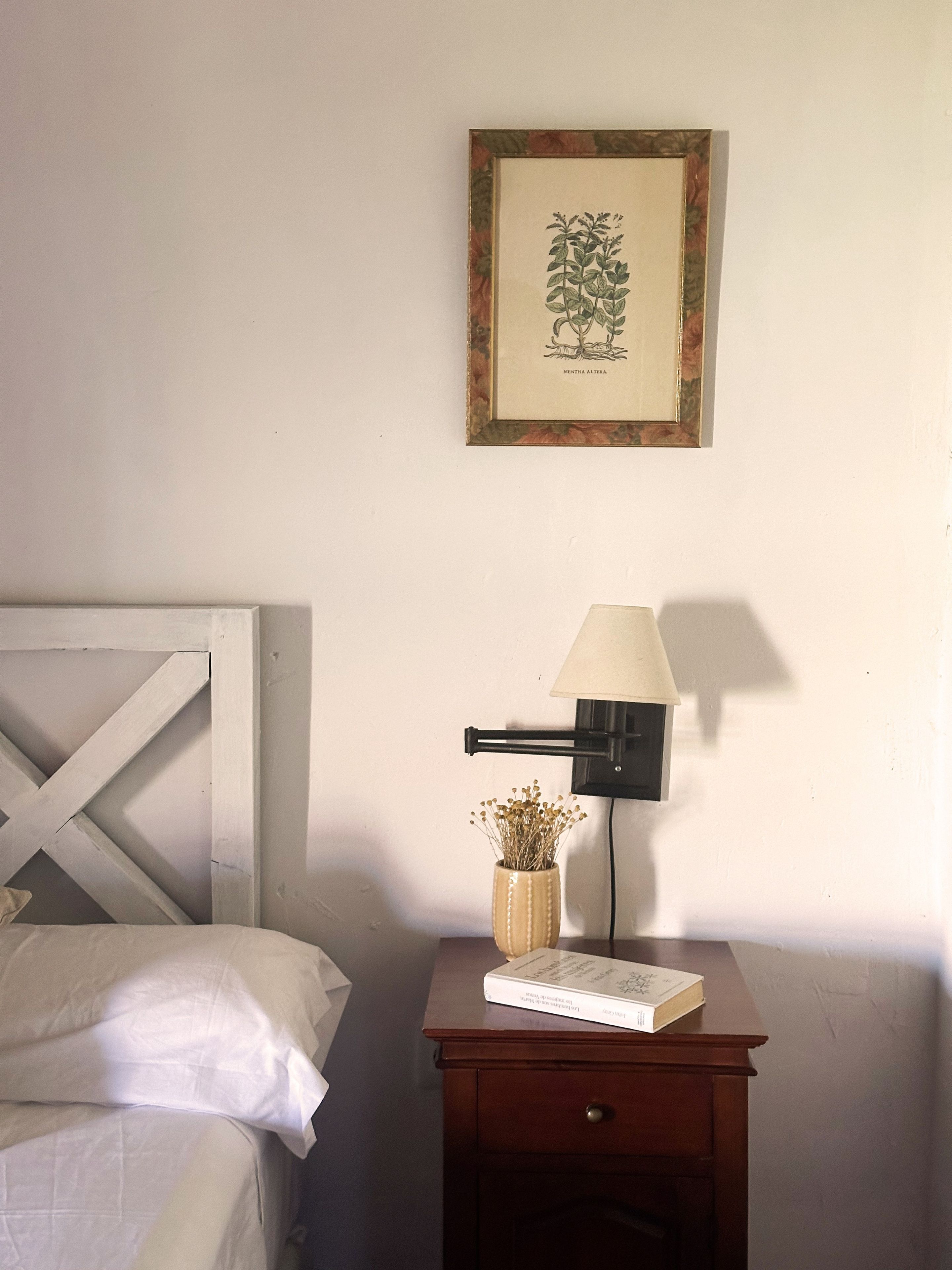 Un dormitorio con cama, una mesita de noche con lámpara, una ilustración botánica enmarcada y un pequeño jarrón con flores secas.