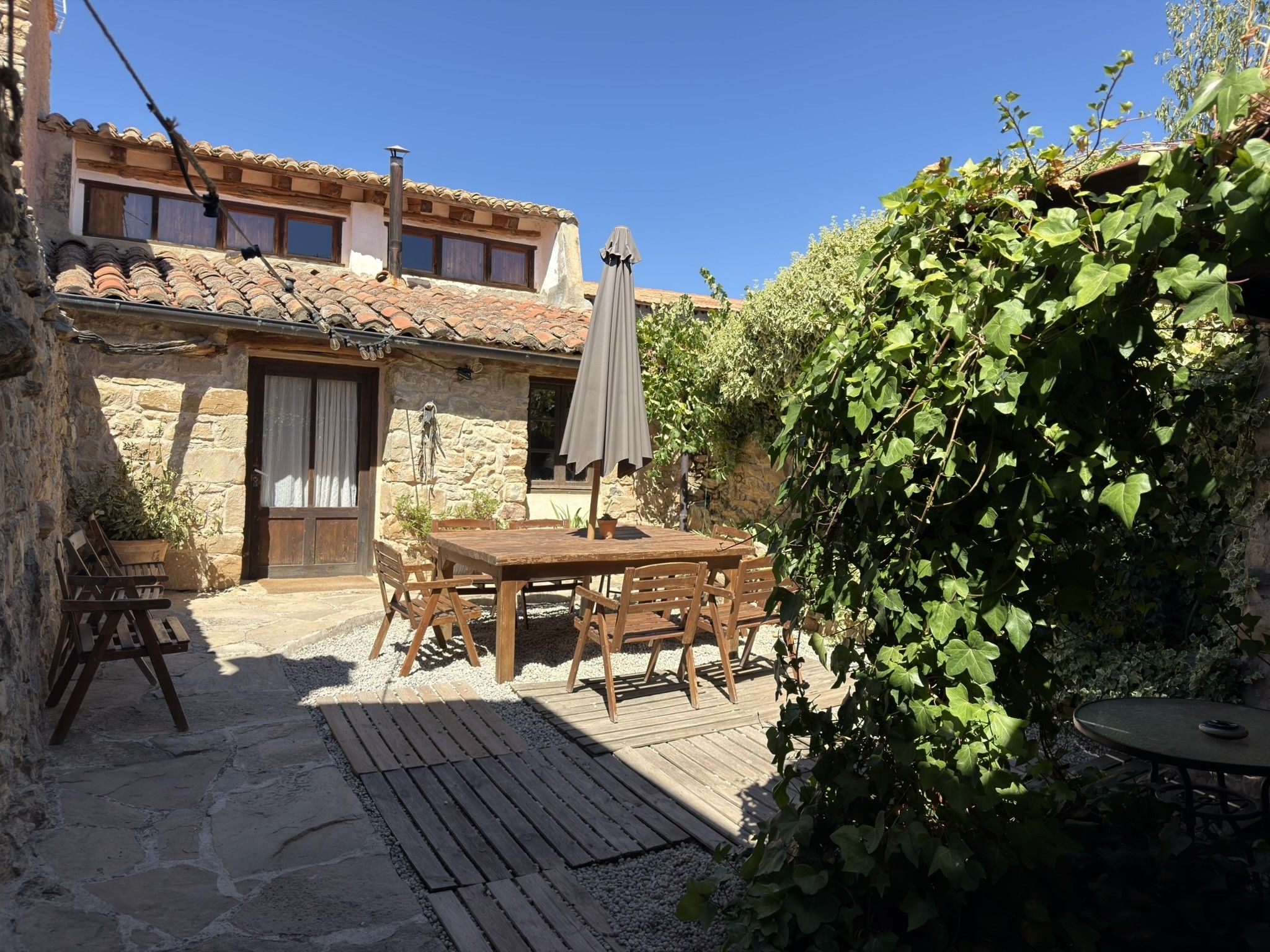 Ein Gebäude aus Stein mit einem Ziegeldach, einem Holztisch mit Stühlen und einer Pergola mit Dach.