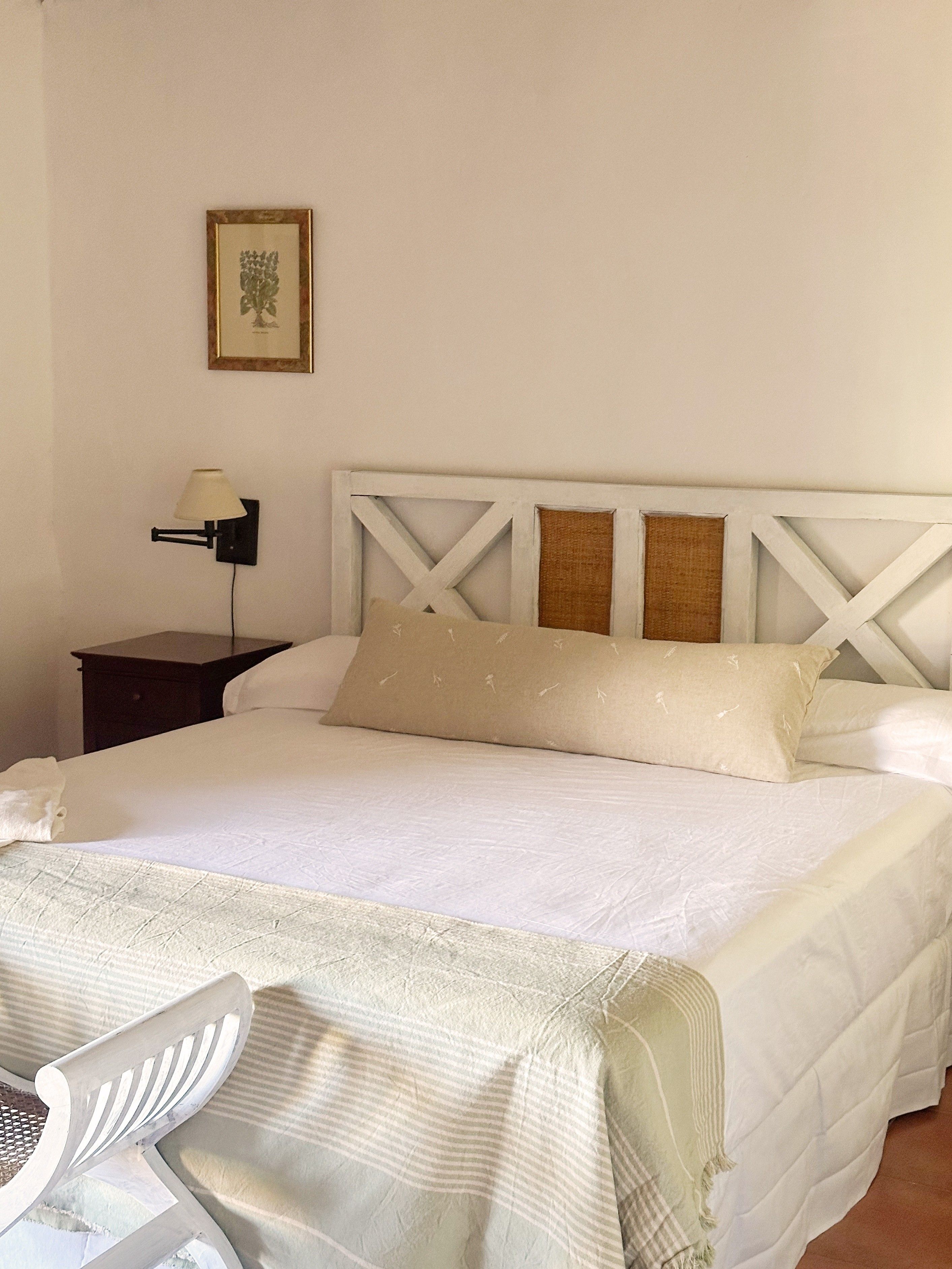 Una cama con cabecera blanca, una mesita de noche con lámpara y una silla.
