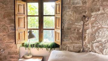 Una habitación rústica con vigas de madera a la vista, una pared de piedra, una ventana con contraventanas de madera, una pequeña mesa con lámpara y un sofá con cojines.