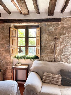Une pièce rustique avec des poutres apparentes, un mur en pierre, une fenêtre avec des volets en bois, une petite table avec une lampe et un canapé avec des coussins.