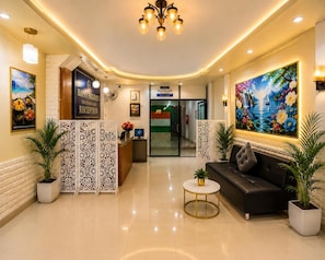 A hotel reception area with a reception desk, a sofa, potted plants, a painting, and a chandelier. - Happy Holidays Hotel (Patong)