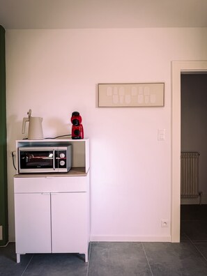 Un coin cuisine équipé d’un micro-ondes, d’une bouilloire et d’une machine à café, le tout posé sur un meuble blanc. On aperçoit également un interrupteur mural et un radiateur à l’arrière-plan.
