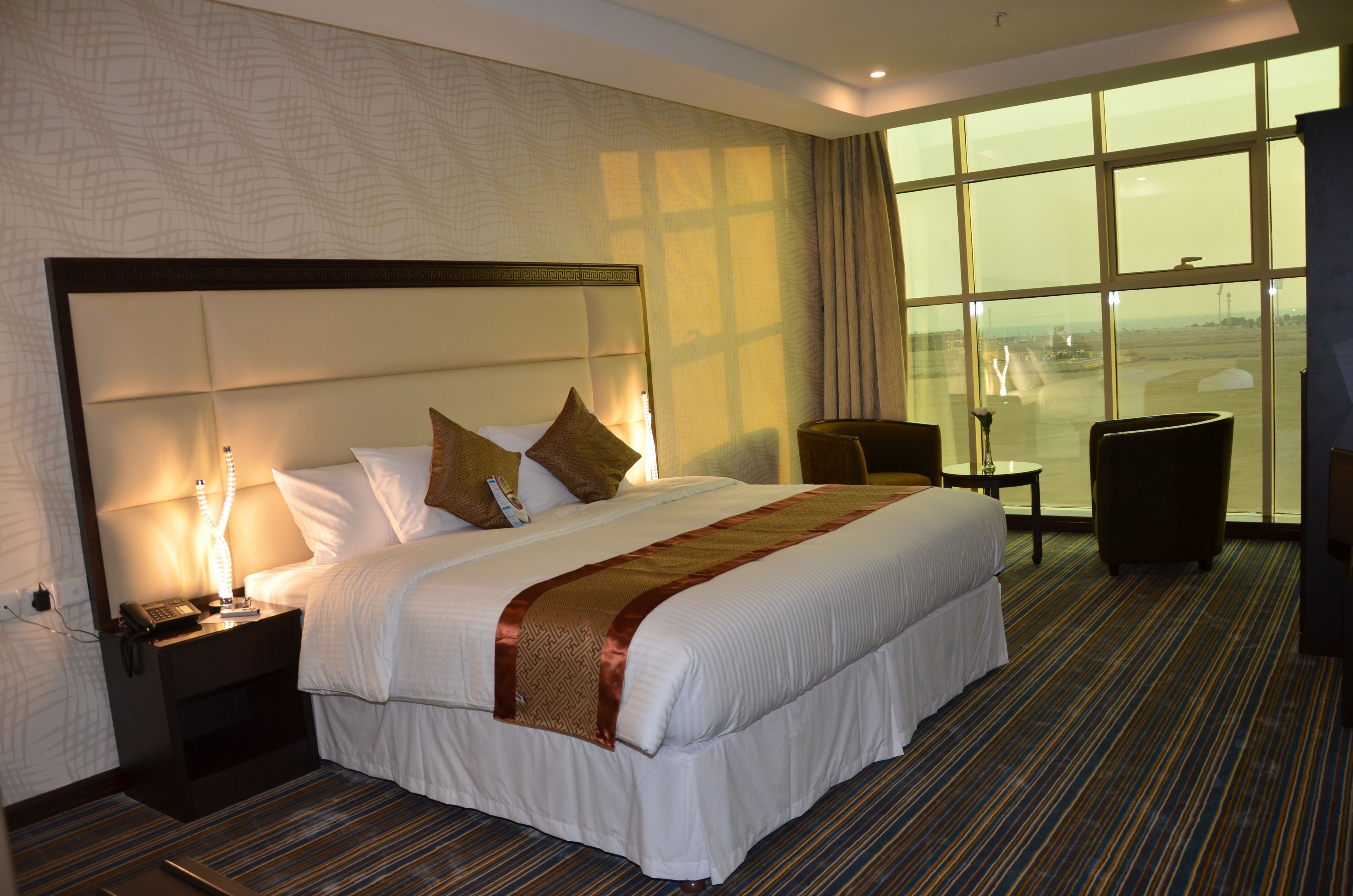 Habitación de hotel con una cama grande, una mesita de noche con lámpara, una silla y vista al aeropuerto.