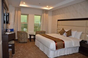 A hotel room with a large bed, a desk, a chair, a small table, and a window with curtains. - Golden New Armada (Jizan)