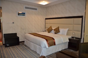 A hotel room with a large bed, a bedside table, a chair, a carpeted floor, a wall-mounted air vent, and a framed picture. - Golden New Armada (Jizan)