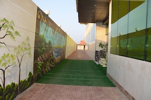 A corridor with a mural on the left wall, a green artificial turf path, and a building with large windows on the right. - Golden New Armada (Jizan)