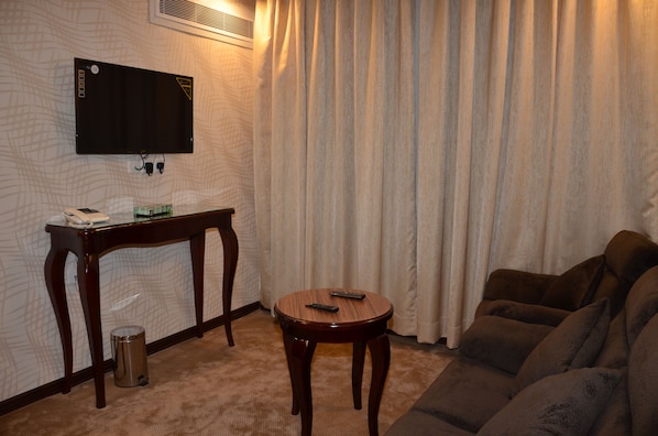 A living room with a flat-screen TV mounted on the wall, a small table with a telephone and a remote control, a trash can, and a sofa. - Golden New Armada (Jizan)