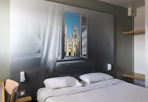 Un dormitorio con una cama, dos almohadas, una mesita de noche, una lámpara de pared y una ventana con vista a una catedral.