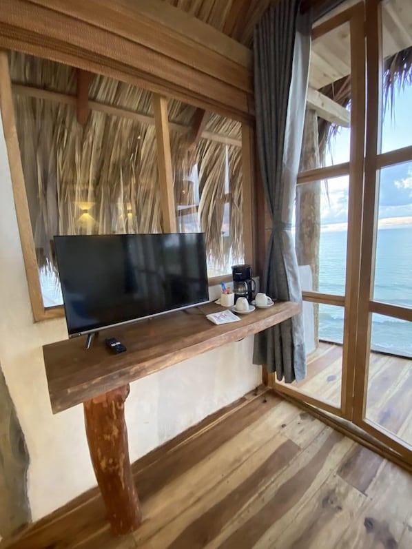 Una habitación con estructura de madera, un ventanal grande con vista al mar, un televisor de pantalla plana sobre un soporte de madera, y una pequeña mesa con una cafetera y tazas.