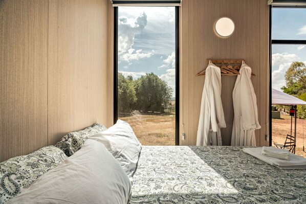 A bedroom with a large bed, a view of the outdoors, and a wooden wardrobe with hanging clothes.