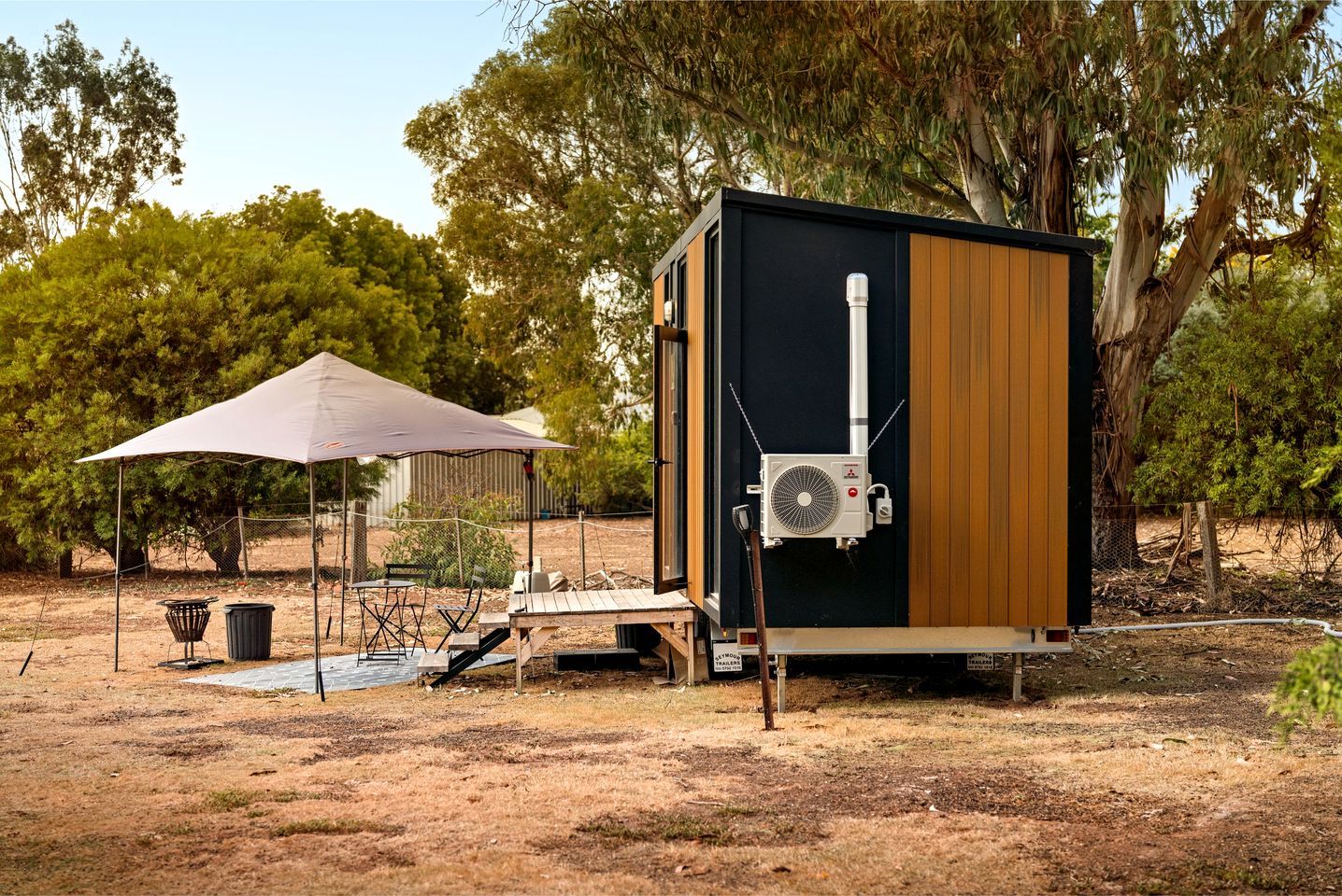 A portable RV with a black and brown exterior, a white air conditioning unit mounted on the side, and a picnic table with chairs under a white canopy.