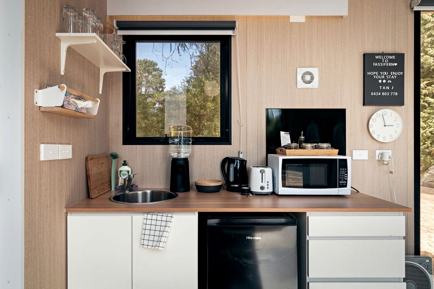 A compact kitchen with a sink, a coffee maker, a toaster, a microwave, and a dishwasher. There is a window with a view of trees, a clock, and a welcome sign.