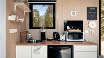 A compact kitchen with a sink, a coffee maker, a toaster, a microwave, and a dishwasher. There is a window with a view of trees, a clock, and a welcome sign.
