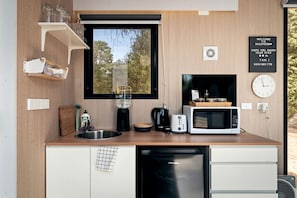 Cocina compacta con lavamanos, cafetera, tostadora, microondas y lavavajillas. Hay una ventana con vista a árboles, un reloj y un cartel de bienvenida.