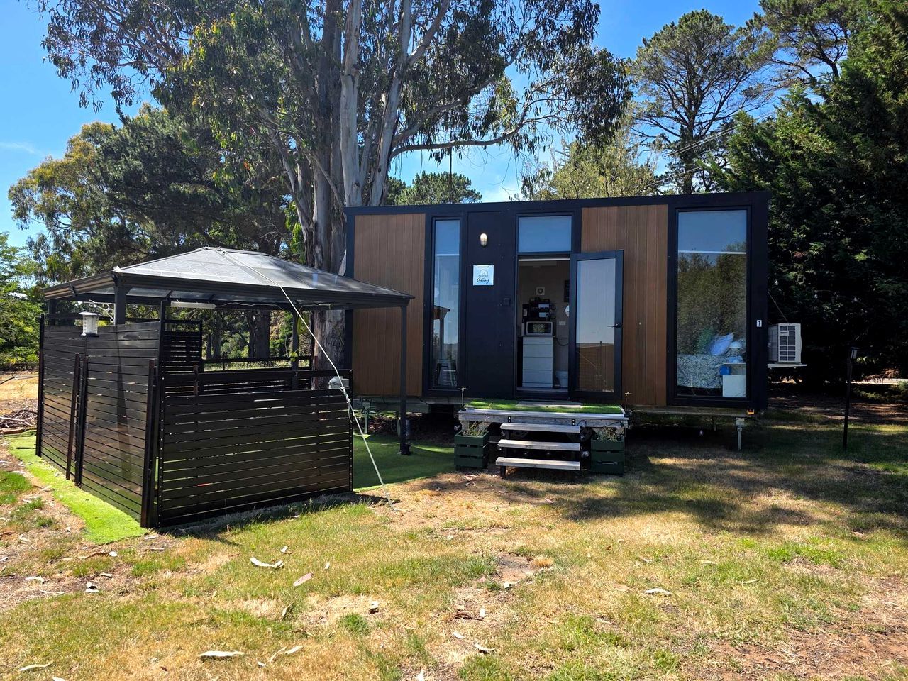 Een modern, compact vakantiehuisje met een houten buitenkant en een glazen voordeur, gelegen in een grasrijke omgeving met bomen op de achtergrond.