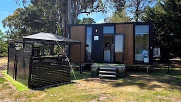 Een modern, compact vakantiehuisje met een houten buitenkant en een glazen voordeur, gelegen in een grasrijke omgeving met bomen op de achtergrond.