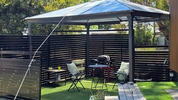 A covered outdoor seating area with a table and chairs, a grill, and a fenced garden.