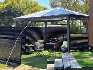 A covered outdoor seating area with a table and chairs, a grill, and a fenced garden.