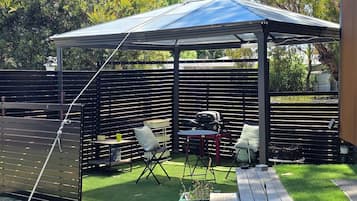 Een overdekt terras met een tafel, stoelen, een barbecue en een omheinde tuin.
