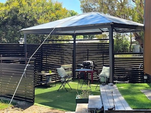 A covered outdoor seating area with a table and chairs, a grill, and a fenced garden.