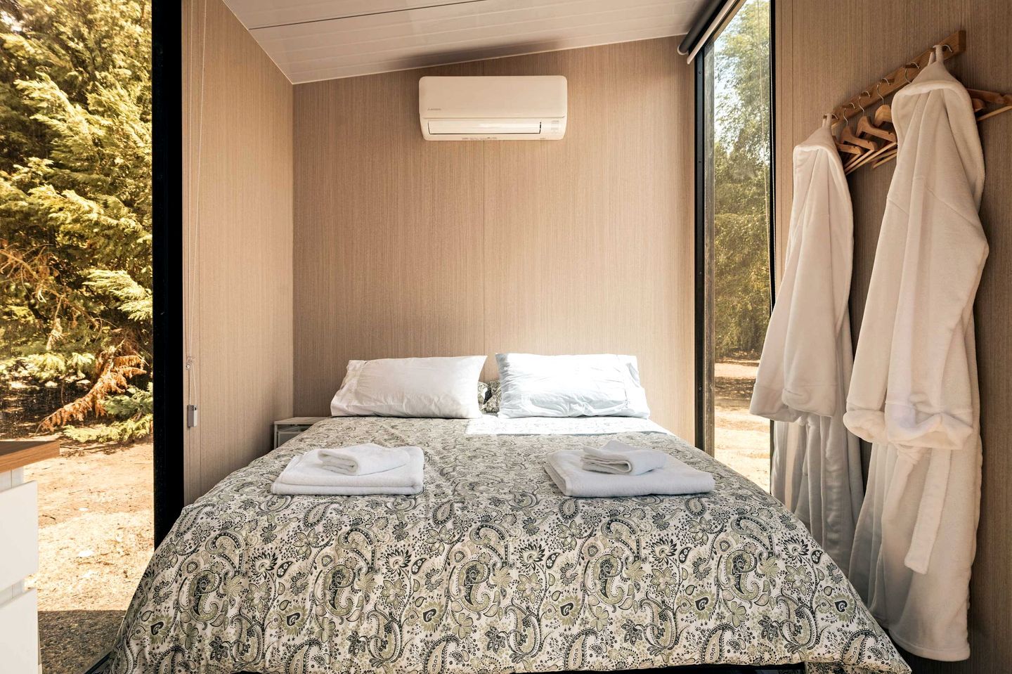 A bedroom with a bed, two pillows, a bedside table with towels, an air conditioning unit, and a view of trees through a window.