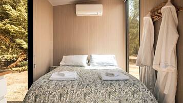 A bedroom with a bed, two pillows, a bedside table with towels, an air conditioning unit, and a view of trees through a window.