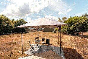 An outdoor patio with a table and chairs under a large umbrella, surrounded by a fenced area with trees and a building in the background.