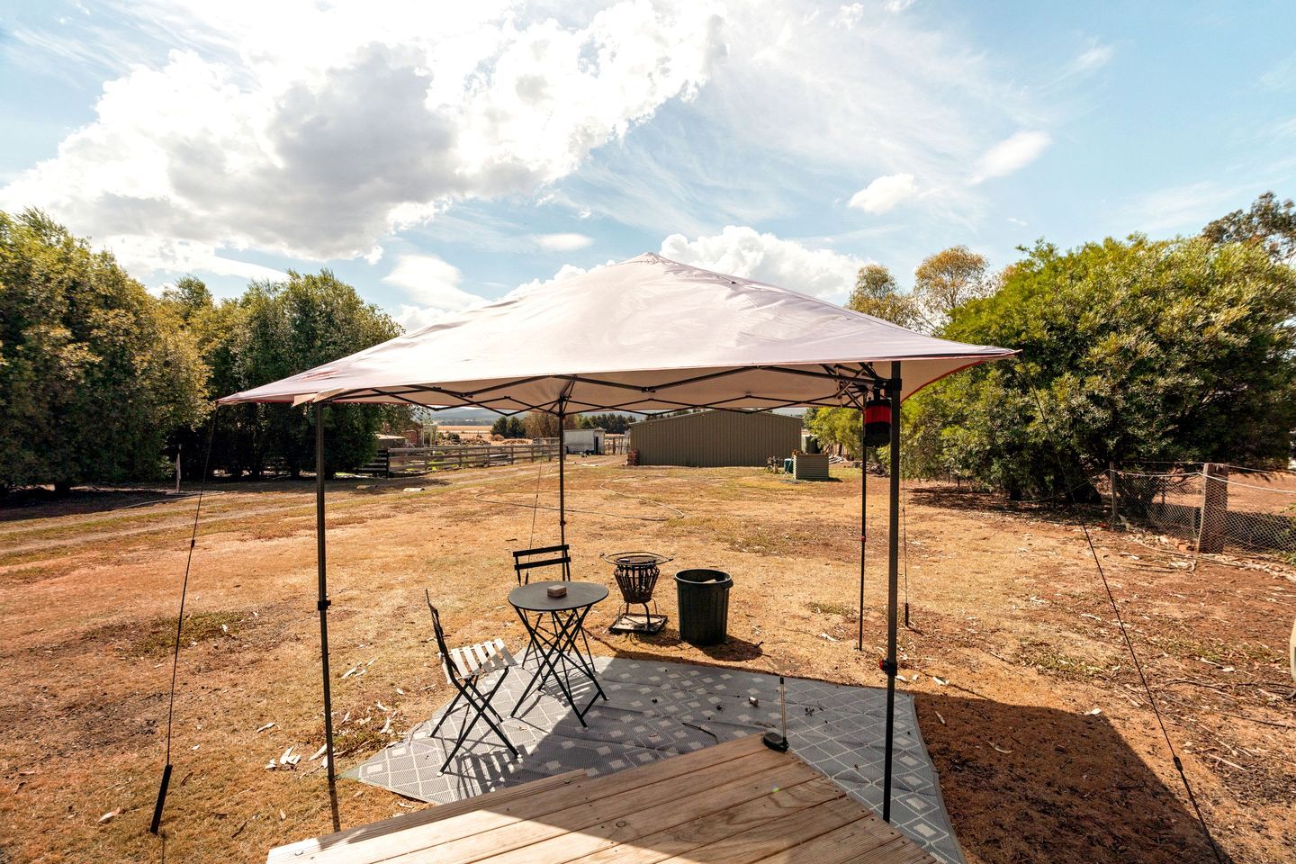 An outdoor patio with a table and chairs under a large umbrella, surrounded by a fenced area with trees and a building in the background.