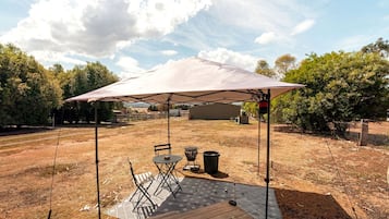 An outdoor patio with a table and chairs under a large umbrella, surrounded by a fenced area with trees and a building in the background.