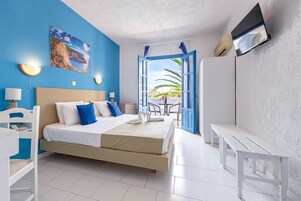 A bedroom with a bed, bedside tables, a desk, and a chair. There is a window with an open door leading to a balcony with a table and chairs.