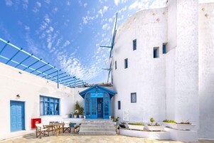Un edificio blanco con puerta y ventanas azules, un área de estar al aire libre techada, y un cielo despejado.