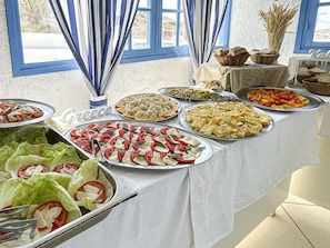 A buffet table with various Greek dishes, a sign reading 'Greece', and a view of the outdoors through a window. - Bluu Bahari Hotel (Karpathos)