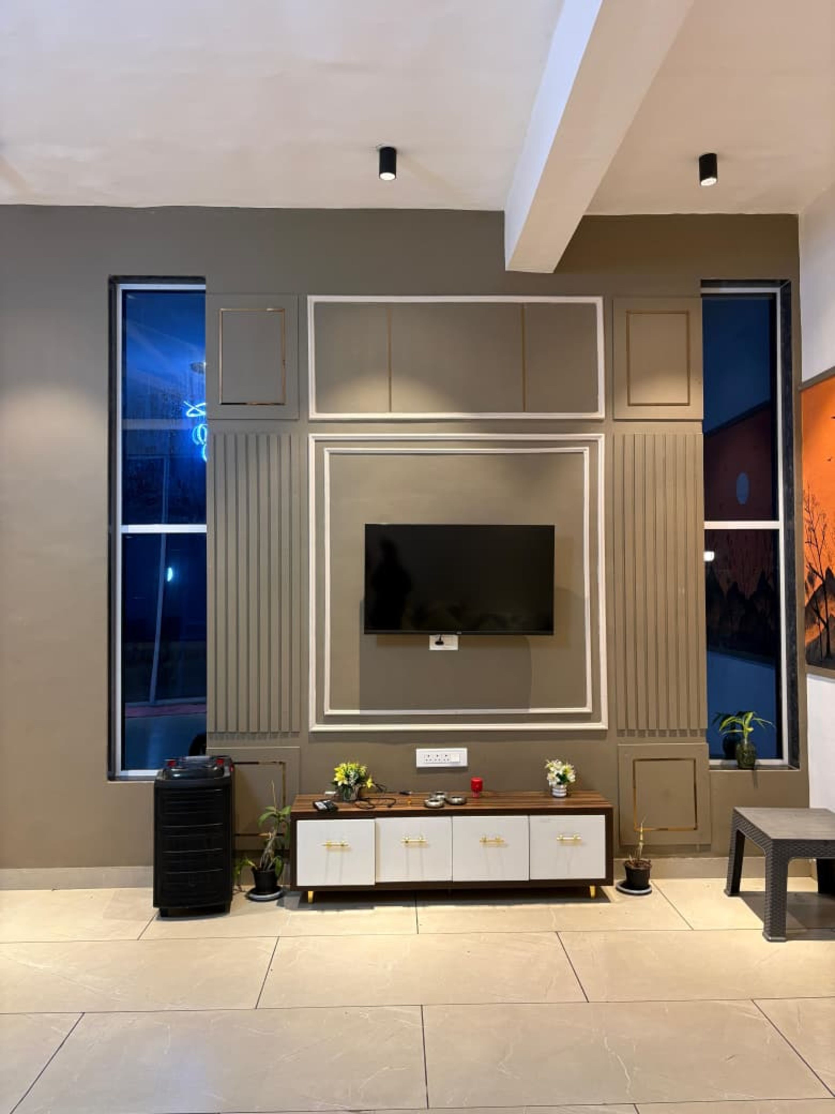 A modern living room with a flat-screen TV mounted on a wall, a large window on one side, and a small table with a potted plant.
