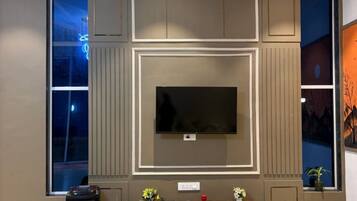 A modern living room with a flat-screen TV mounted on a wall, a large window on one side, and a small table with a potted plant.