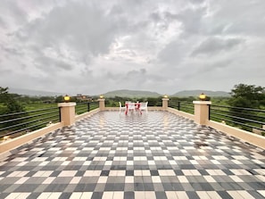 A checkered outdoor terrace with a table and chairs, surrounded by a railing and overlooking a hilly landscape. - Niyati Villa (Chikhalse)