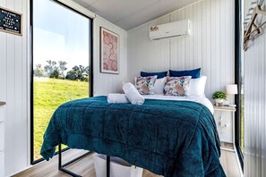 A bedroom with a bed, a window, a wall-mounted air conditioner, and a door. - Charming Tiny Home for a Nature Escape in the Golden Plains Shire near Ballarat, Victoria (Ross Creek)