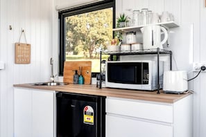 A compact kitchen with a microwave, toaster, and a dishwasher. There is a window with a view of trees and a wooden cutting board with a bottle and a spoon. - Charming Tiny Home for a Nature Escape in the Golden Plains Shire near Ballarat, Victoria (Ross Creek)